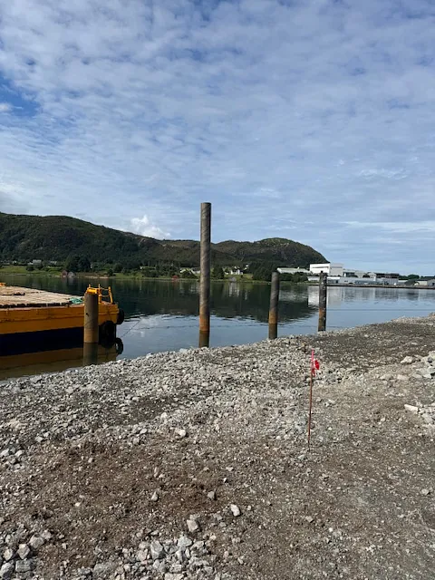 Kaianlegg under bygging med stålpeler ved sjøen, omgitt av fjell og kystlandskap.
